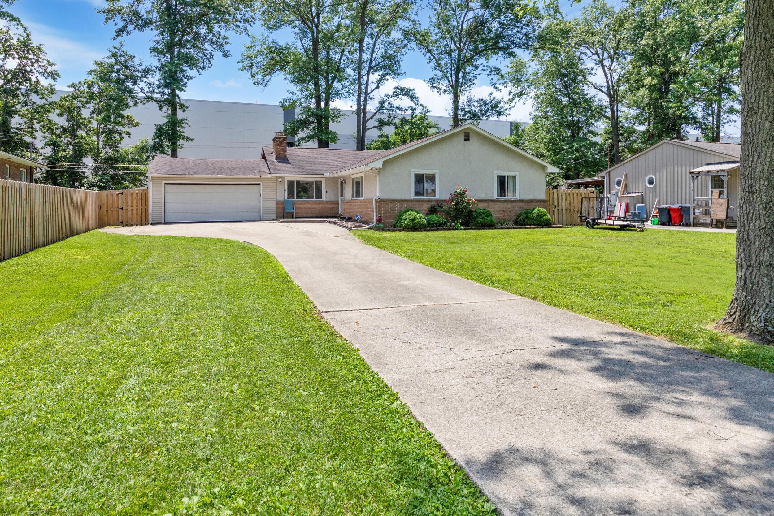 Single-story ranch house with an attached garage, curved driveway, fenced lawn, and mature trees.