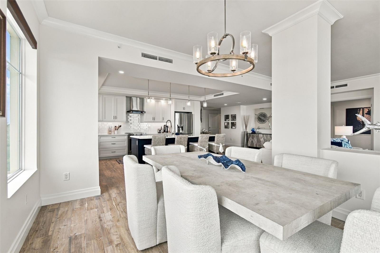 Open-plan dining room with a long pale table and chandelier beside a white kitchen.