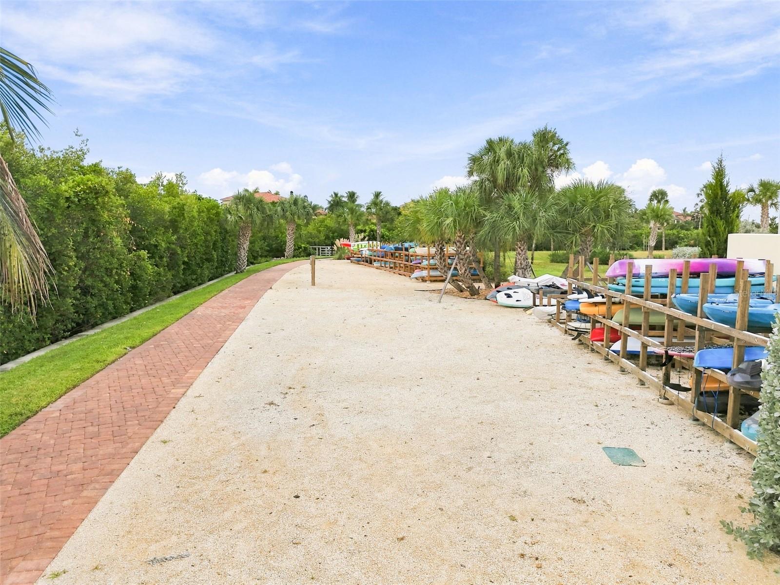 Community kayak and paddleboard storage beside a gravel path and palm trees.