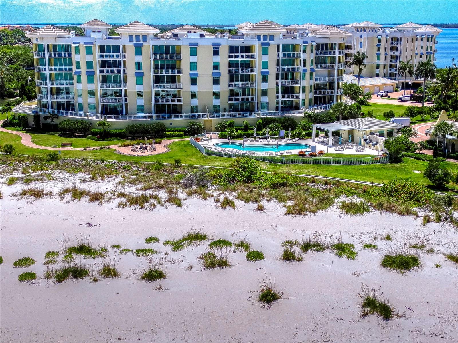 Beachfront condominium buildings with balconies, a courtyard pool, sandy beach, and blue water.