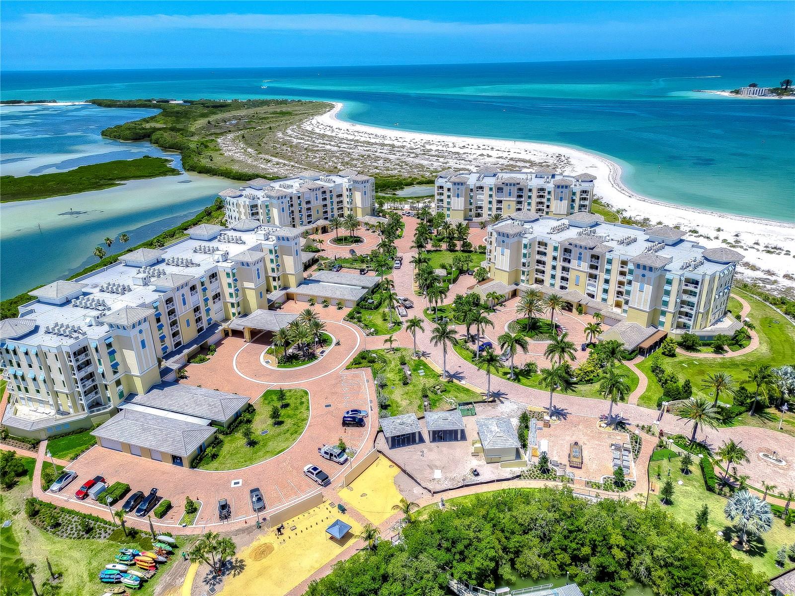 Waterfront condominium complex beside a white-sand beach, viewed from above with turquoise water beyond.