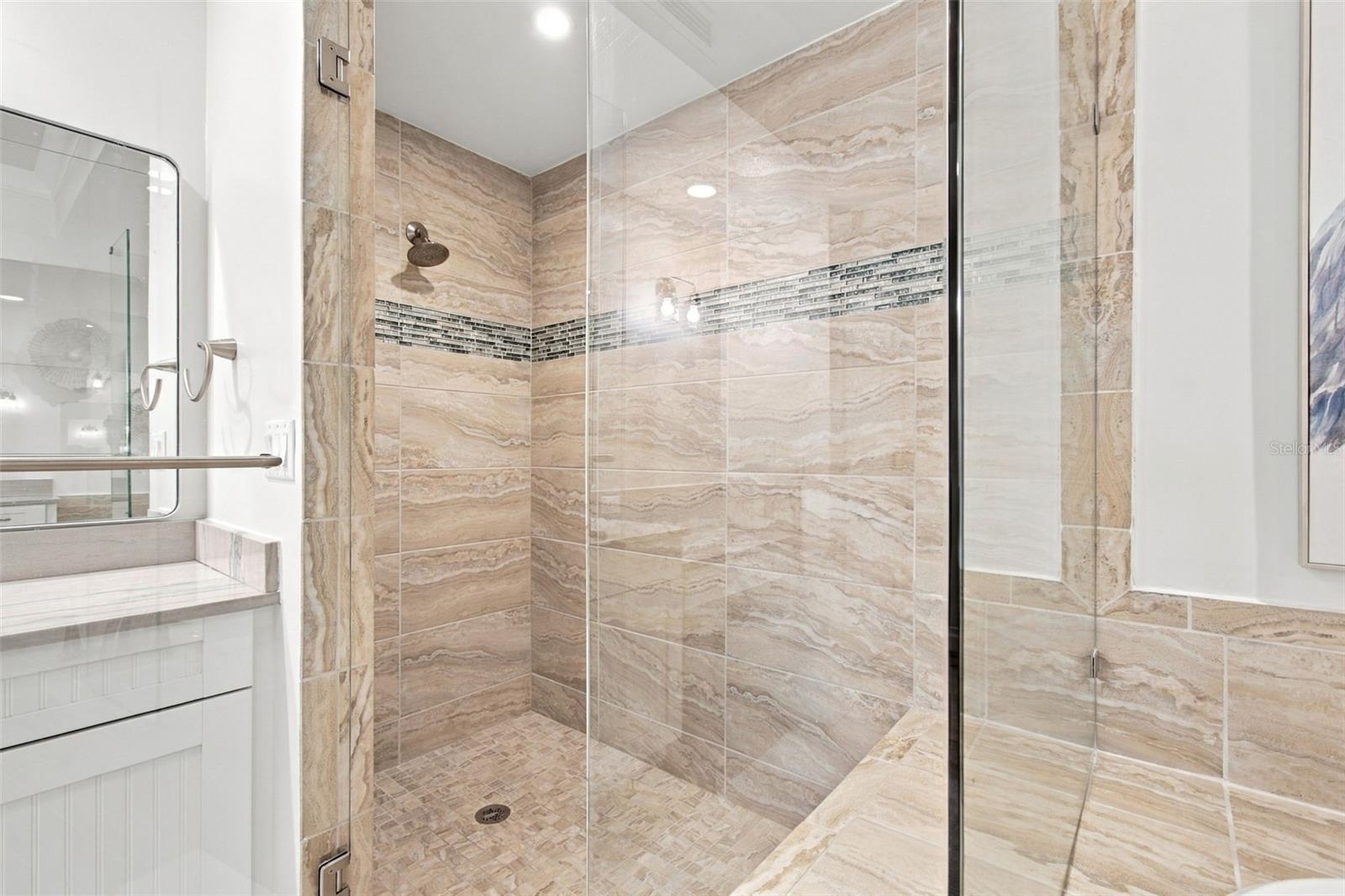 Bathroom with a glass-enclosed, beige-tiled shower and dark mosaic accent stripe beside a white vanity.