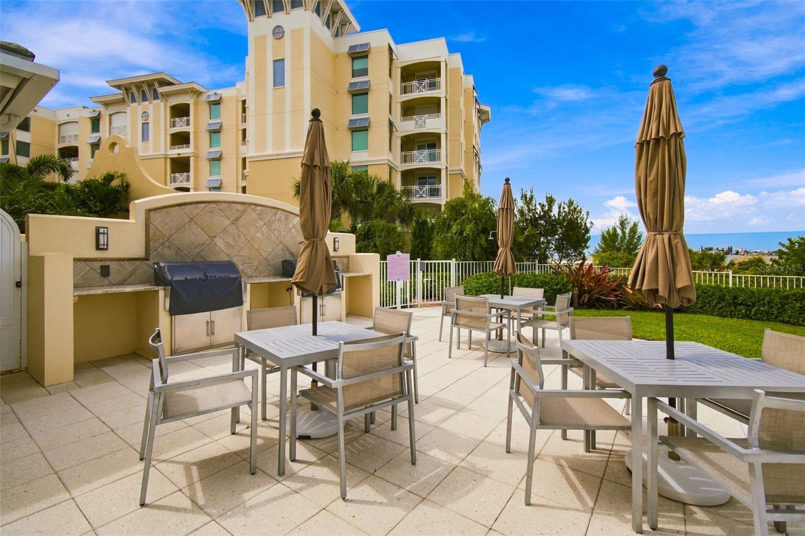 Condominium patio with outdoor dining tables, closed umbrellas, a built-in grill, and a glimpse of water.