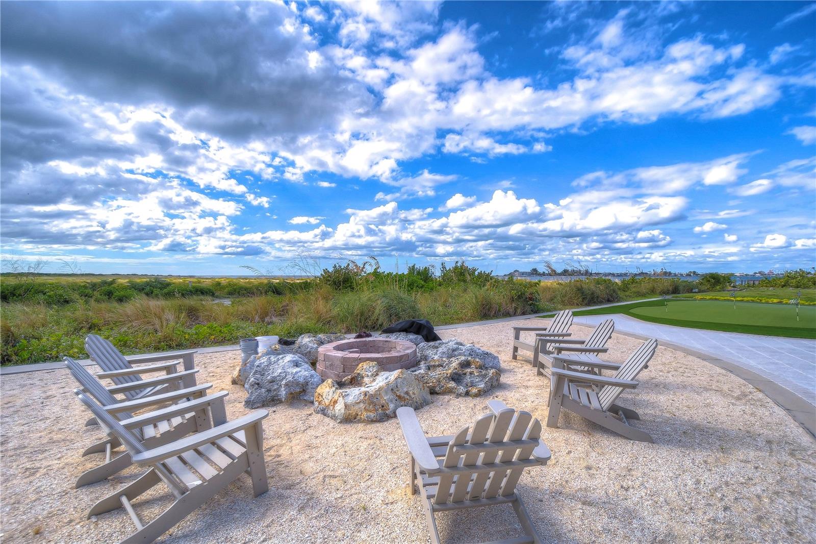 Outdoor seating surrounds a stone fire pit beside a putting green, with coastal vegetation and water in the distance.