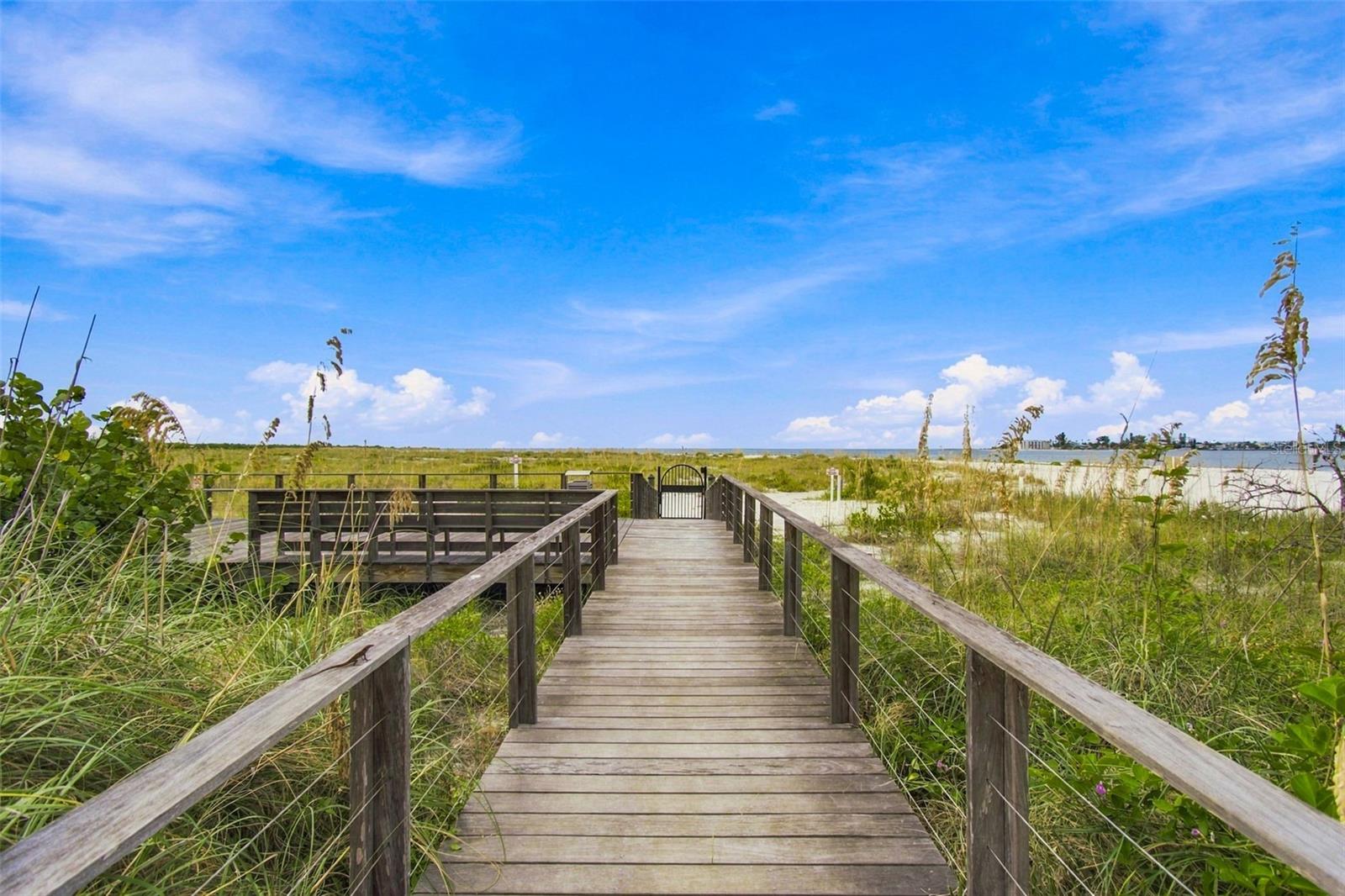 Wooden boardwalk crossing grassy dunes toward beach access and water.