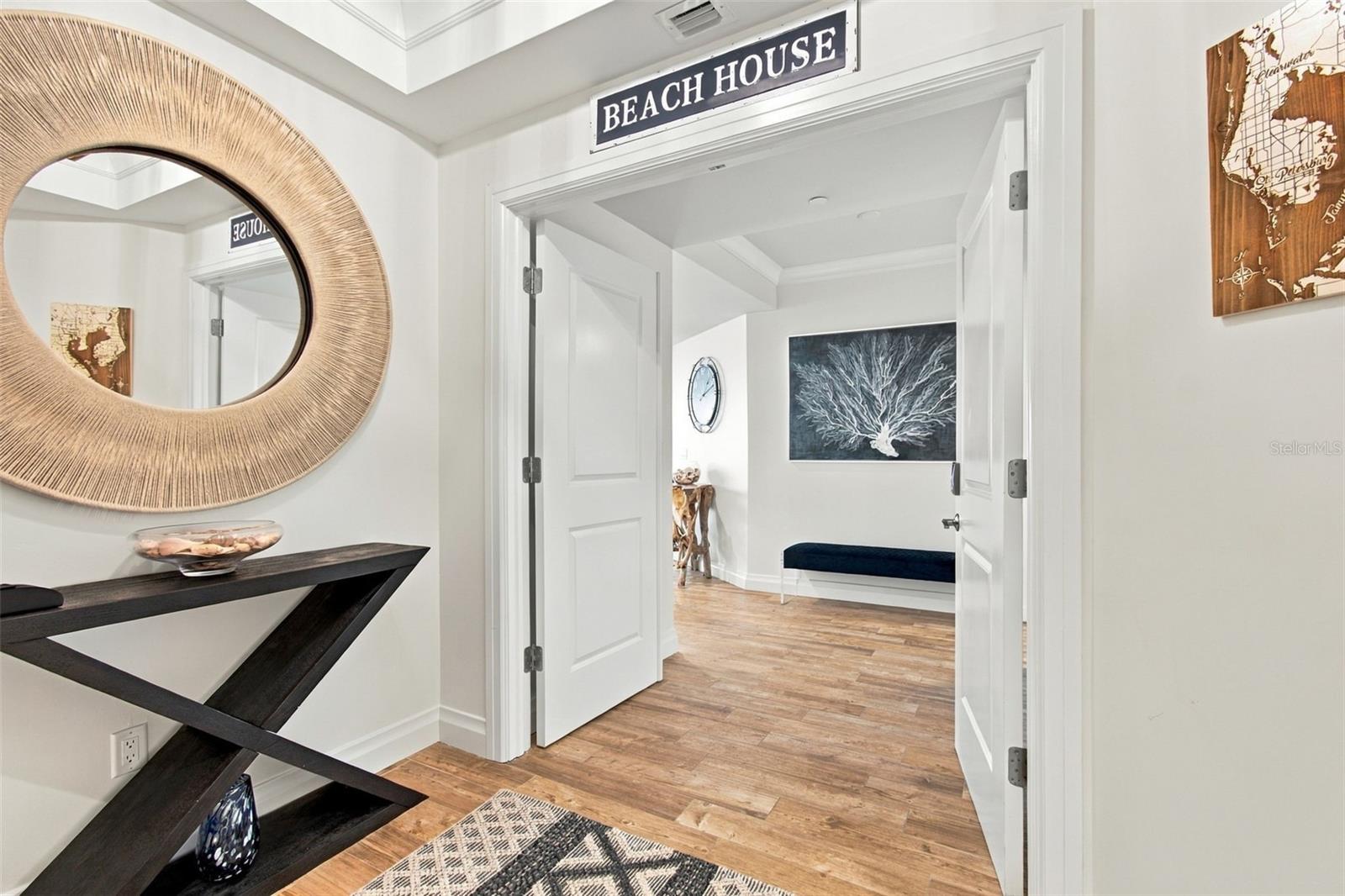 Staged condo entry with a round woven mirror, black console, wood-look floors, and a doorway sign reading “BEACH HOUSE.”
