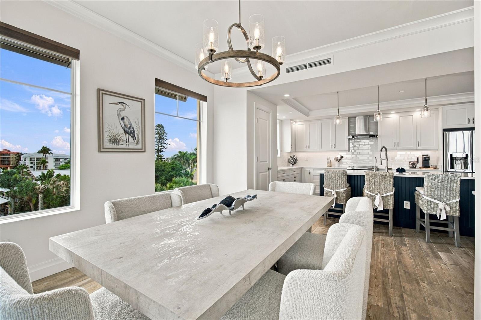 Dining room with a long pale table, opening to a white kitchen with a dark blue island.