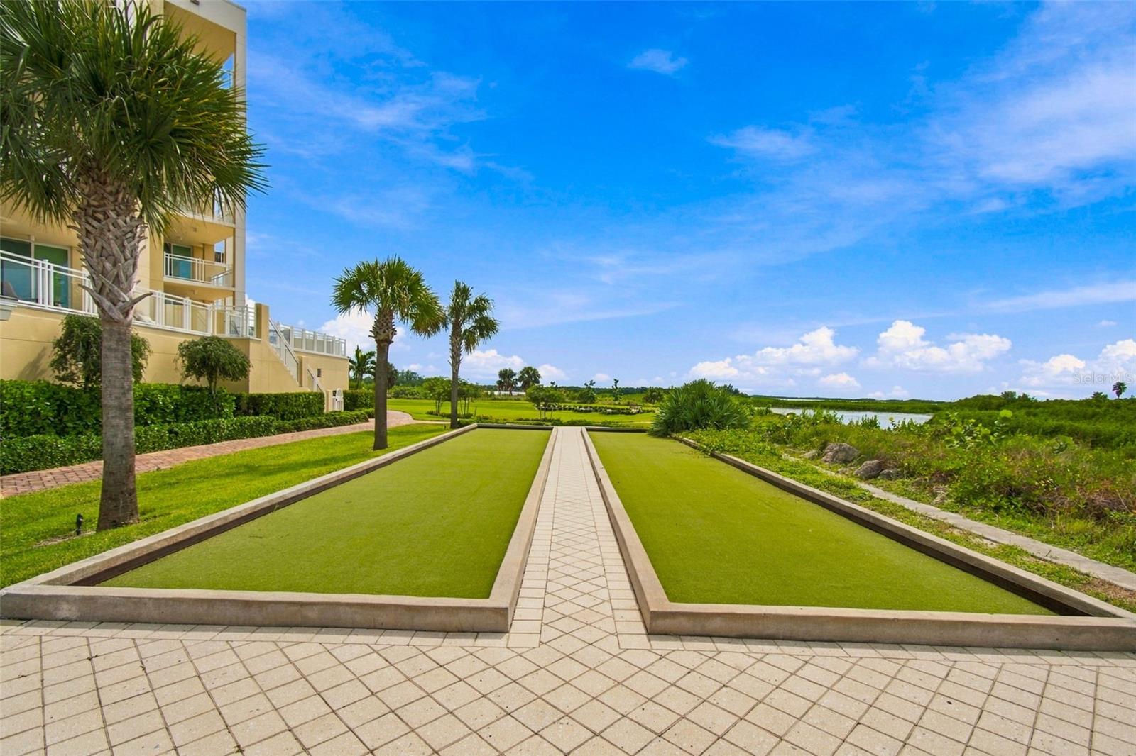 Bocce court beside a condominium, with palm trees and a waterway beyond.