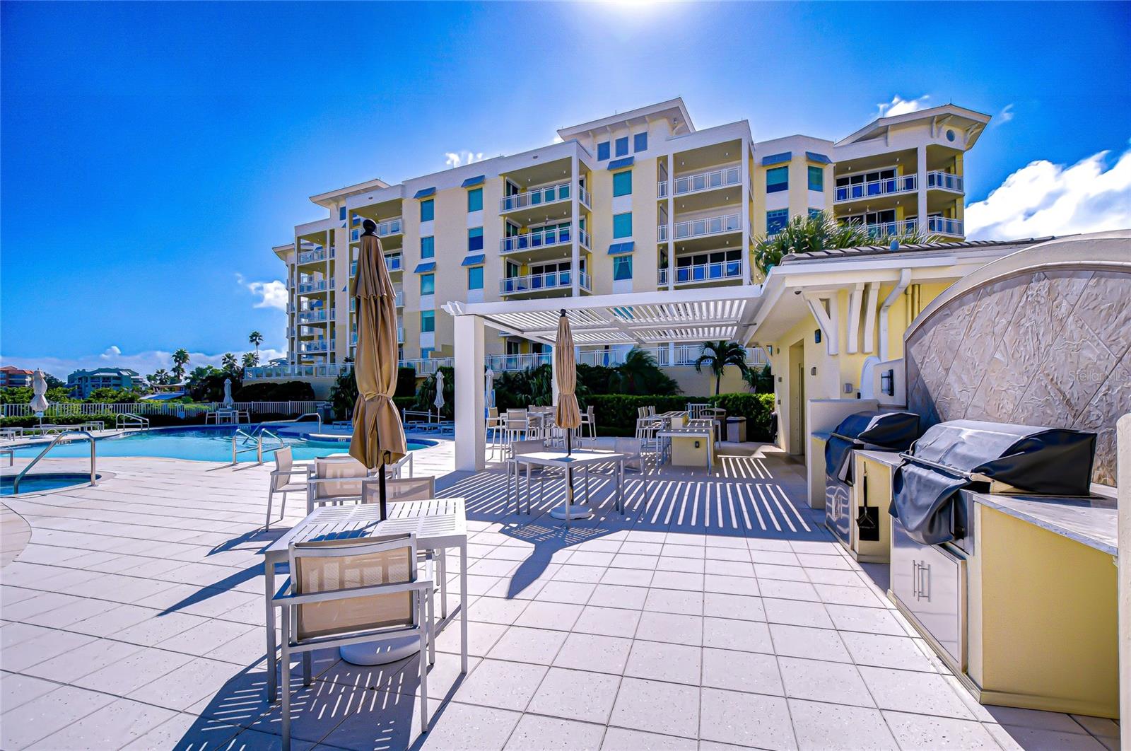 Condominium pool deck with a pergola, outdoor dining furniture, and built-in grill beside the building.