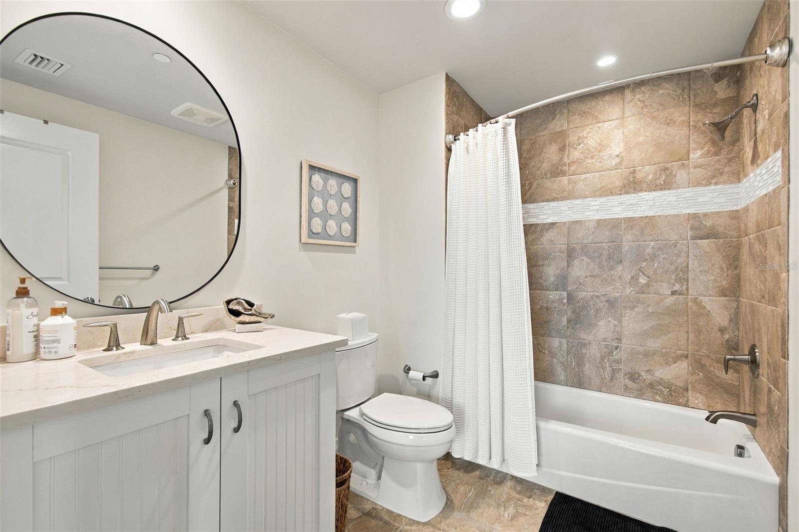 Bathroom with a white vanity and round mirror beside a brown-tiled bathtub and shower.