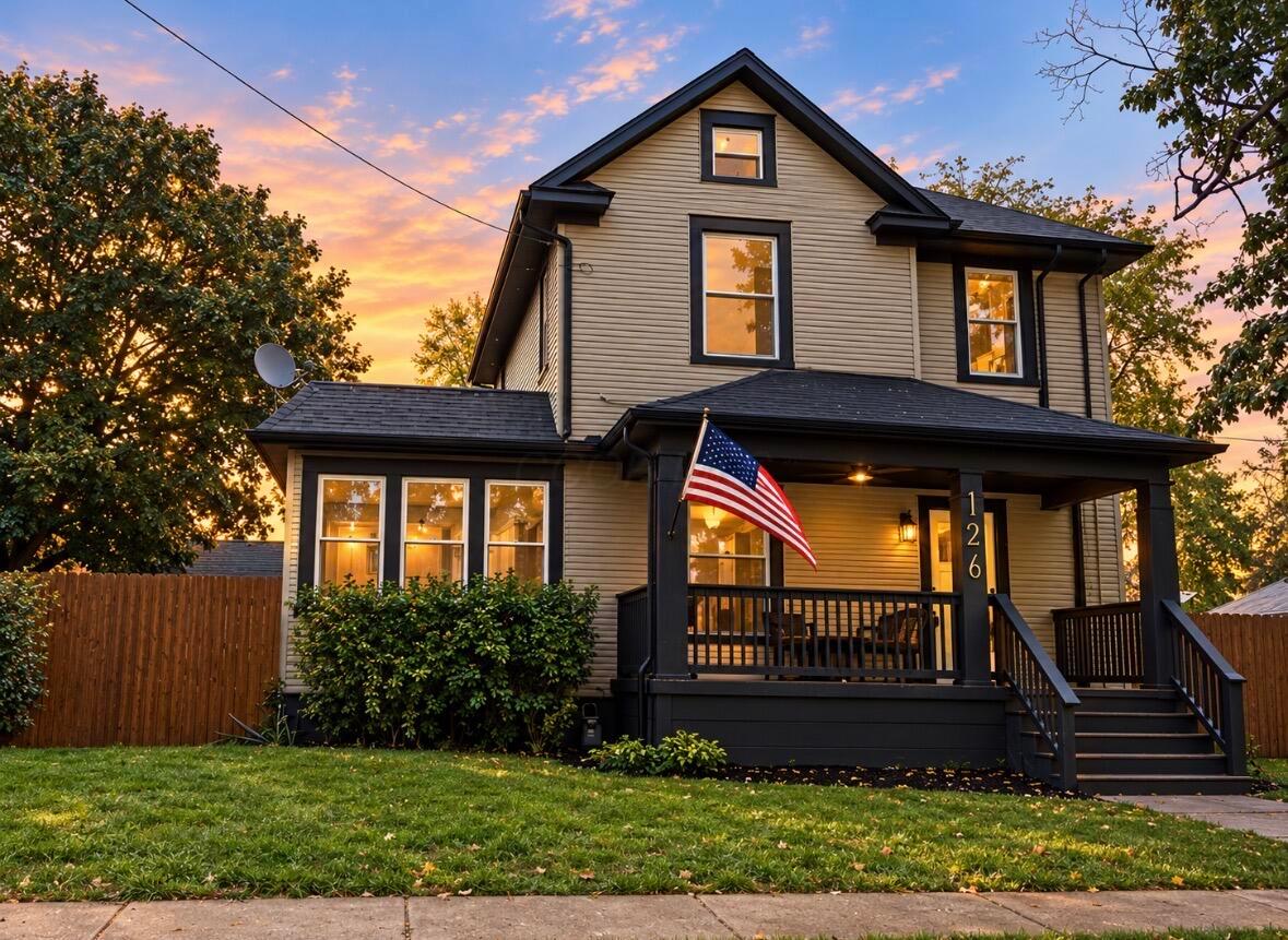 Two-story beige house with a covered porch and American flag at sunset.