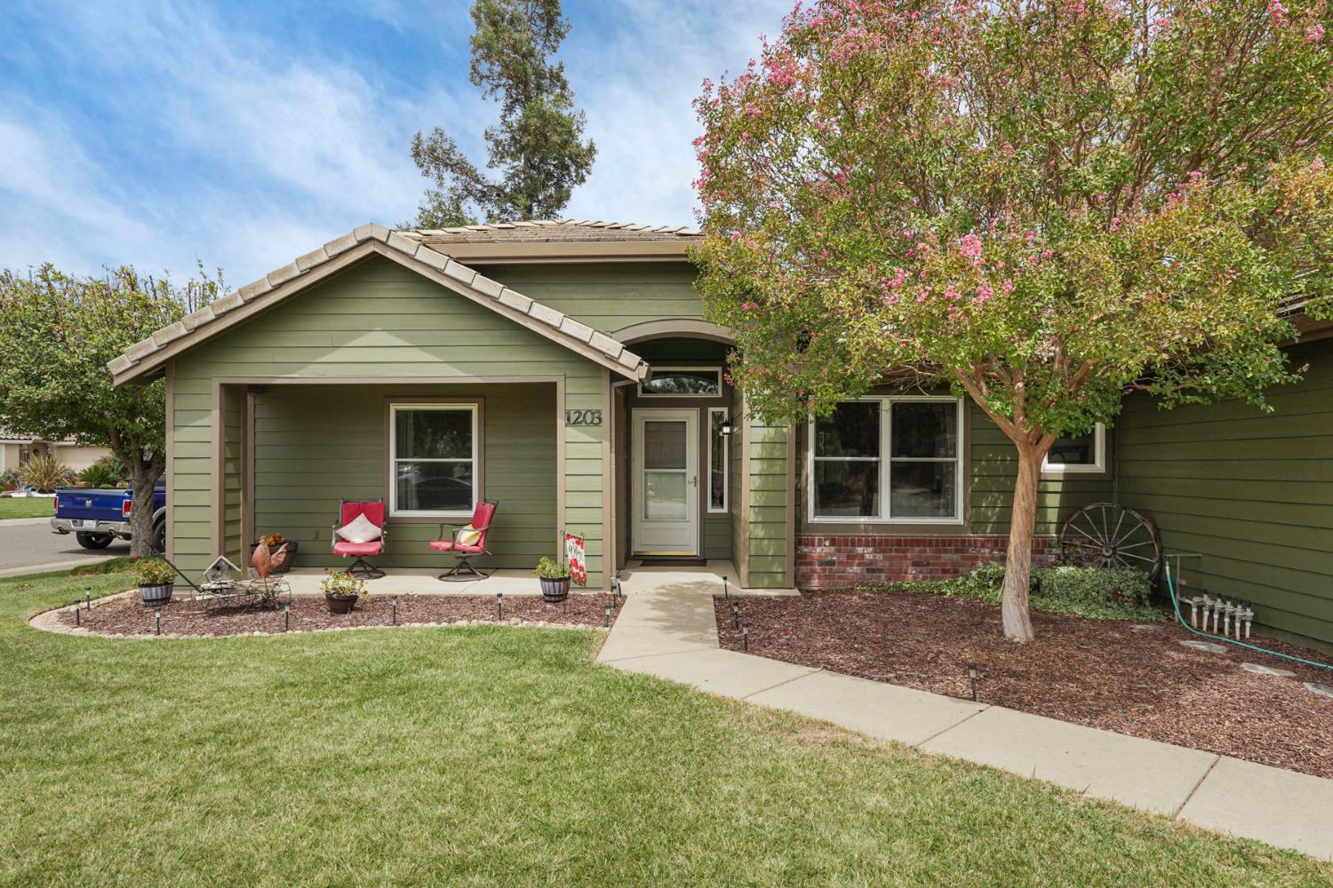 Green-sided home with a covered porch, flowering tree, and curved front walkway.