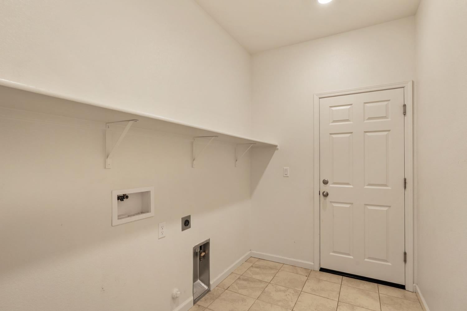 Empty laundry room with wall shelving, washer and dryer hookups, tiled floor, and a paneled door.