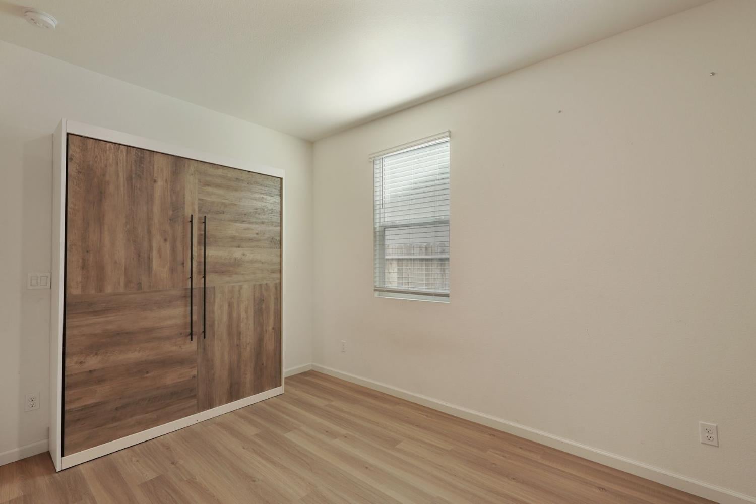 Empty bedroom with a large wood-finished wardrobe, light flooring, and a window with blinds.