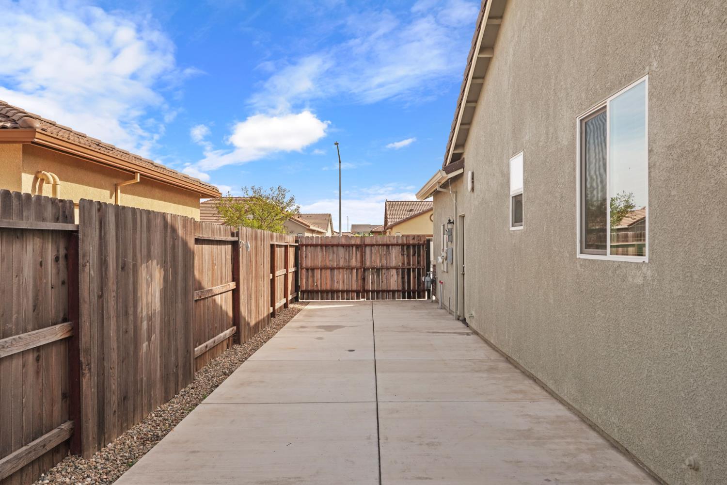 Paved side yard beside a stucco house, enclosed by a tall wooden privacy fence.
