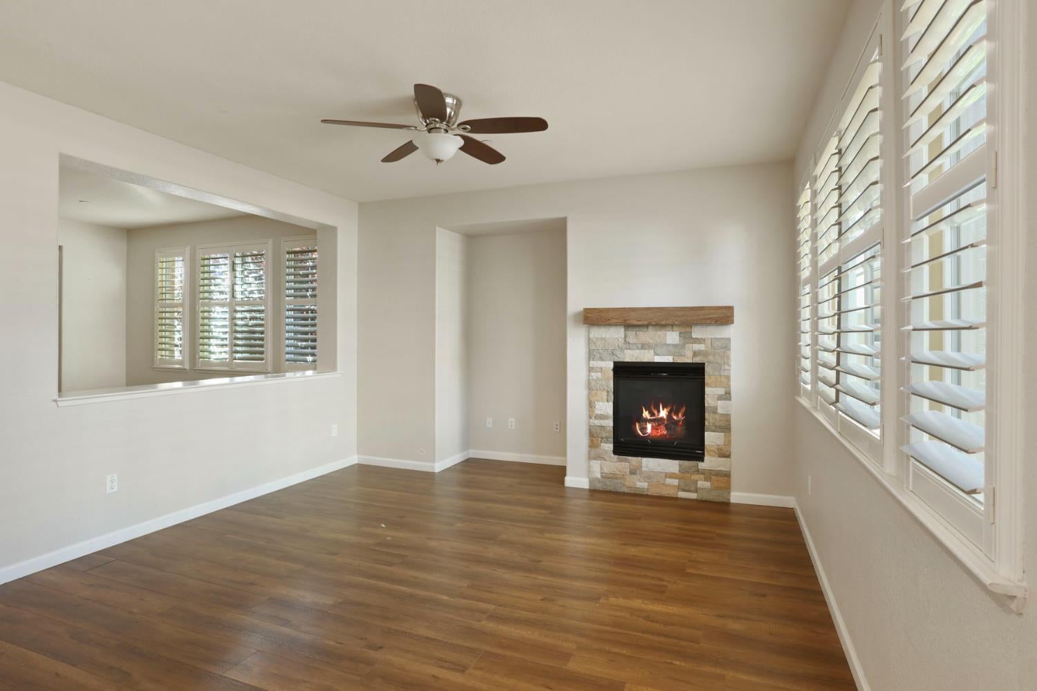 Vacant living room with a stone fireplace, wood floors, and large shuttered windows.