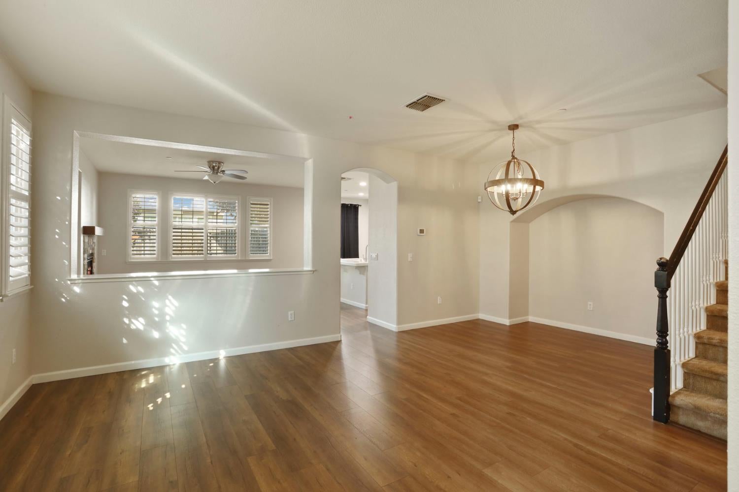 Empty living and dining area with wood floors, a globe pendant, arched niche, and staircase.