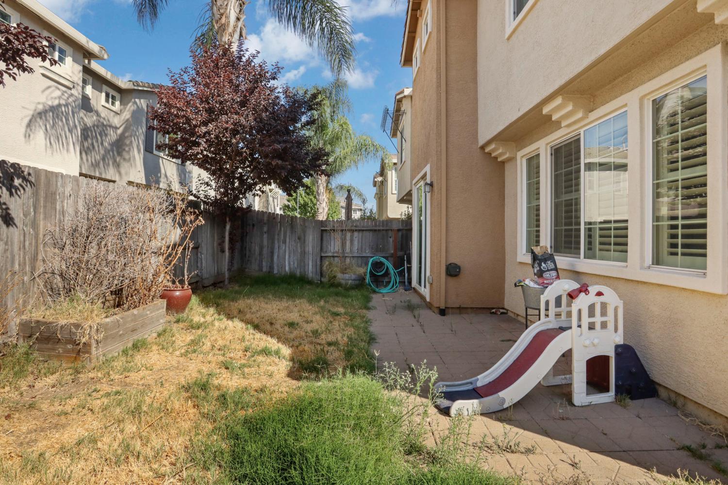 Fenced backyard with a paved patio, raised planter, and children's slide beside a stucco home.