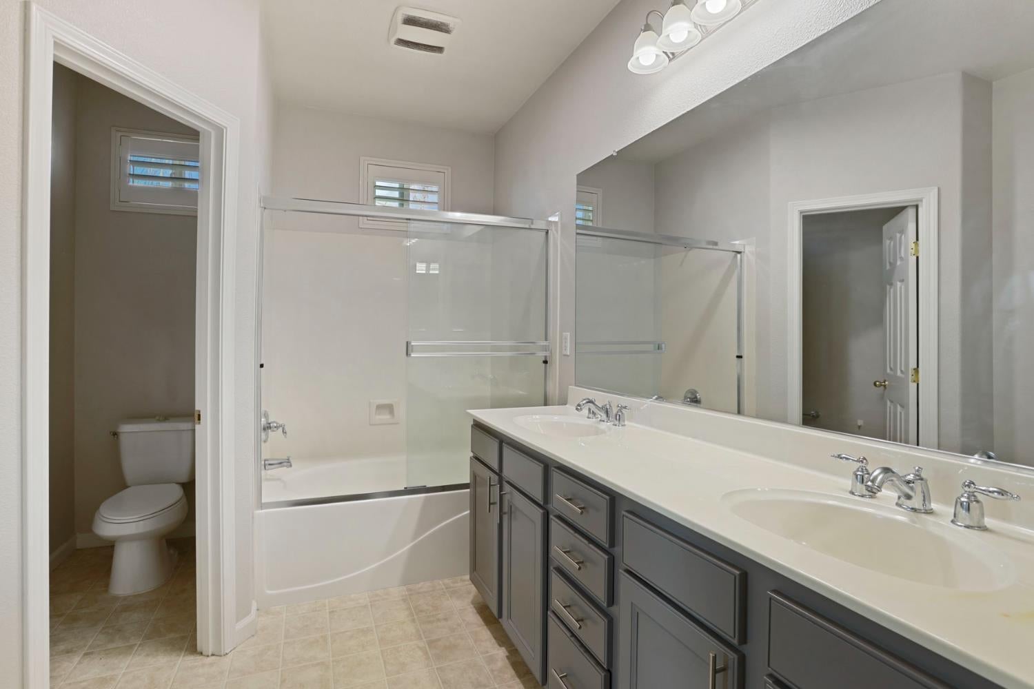 Bathroom with a gray double-sink vanity, large mirror, glass-enclosed bathtub, and separate toilet.
