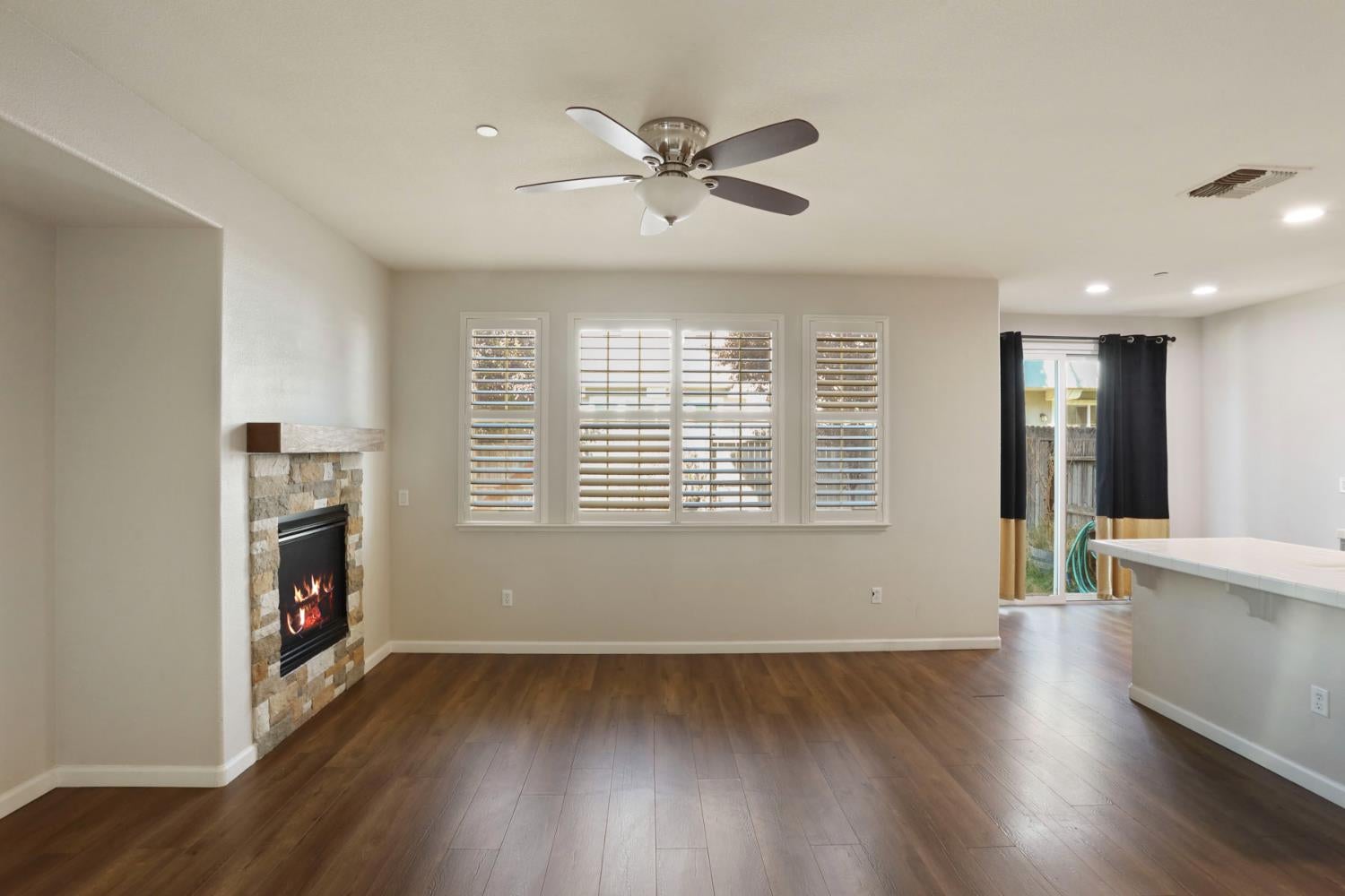 Empty living room with a stone fireplace, plantation shutters, and dark wood floors.
