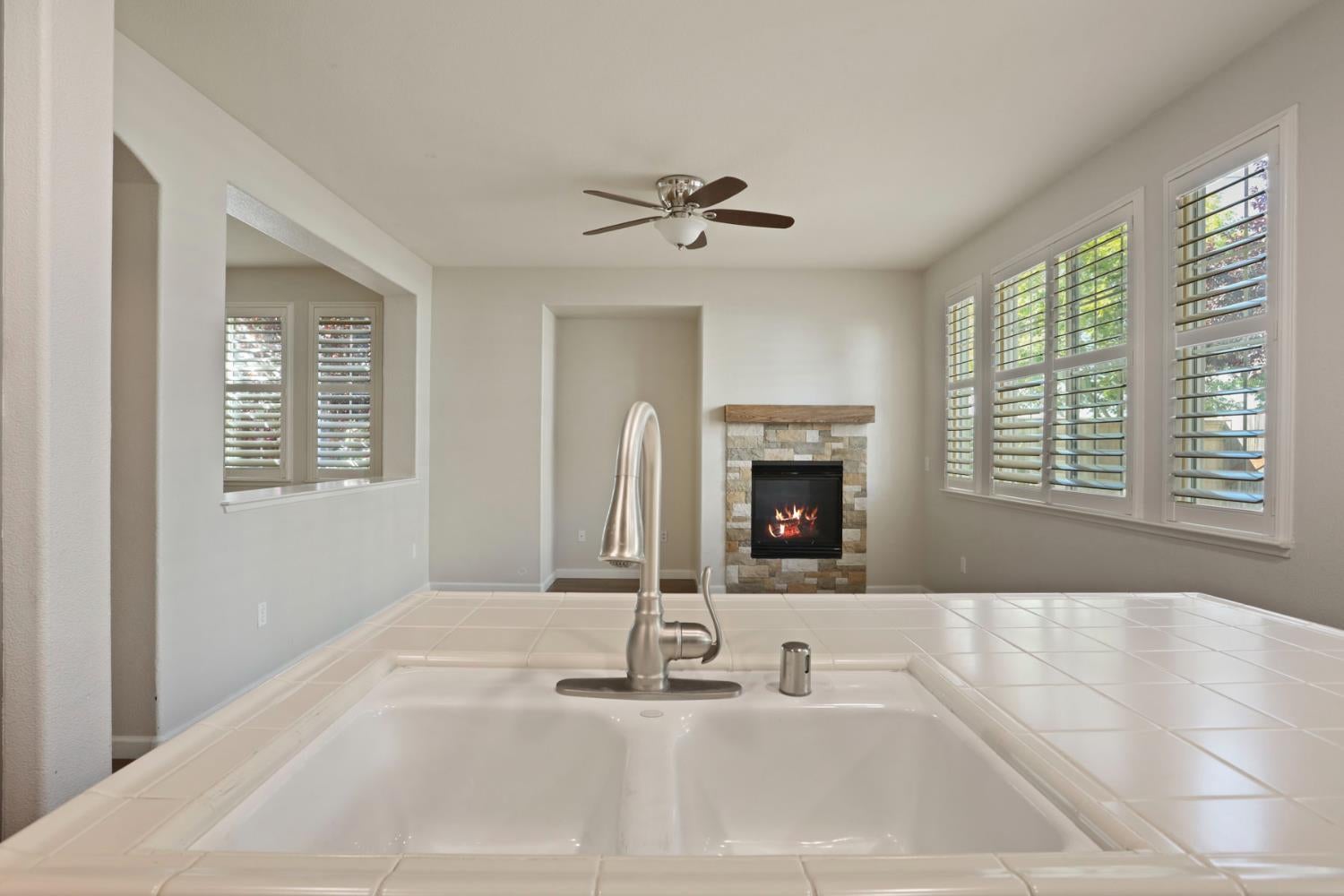 Kitchen sink and tiled counter overlook a living area with a stone fireplace and shuttered windows.