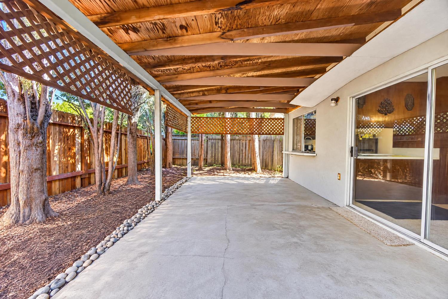 Covered concrete patio with a wooden roof, lattice panels, and trees along the fenced yard.