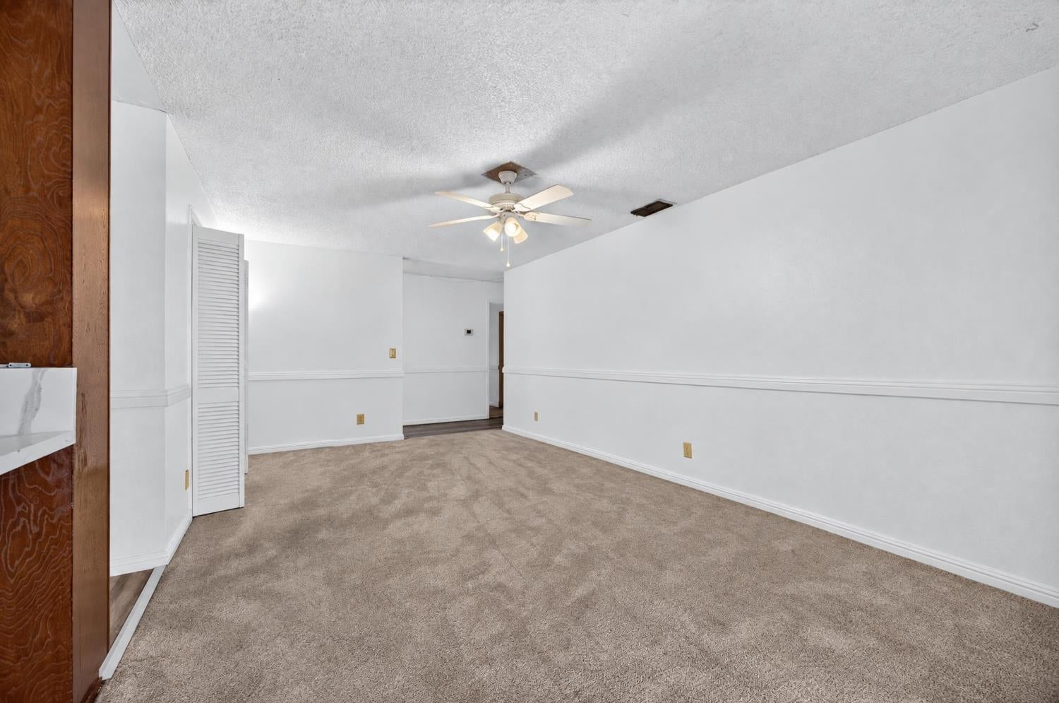 Empty carpeted living room with white walls, a ceiling fan, and a louvered closet door.