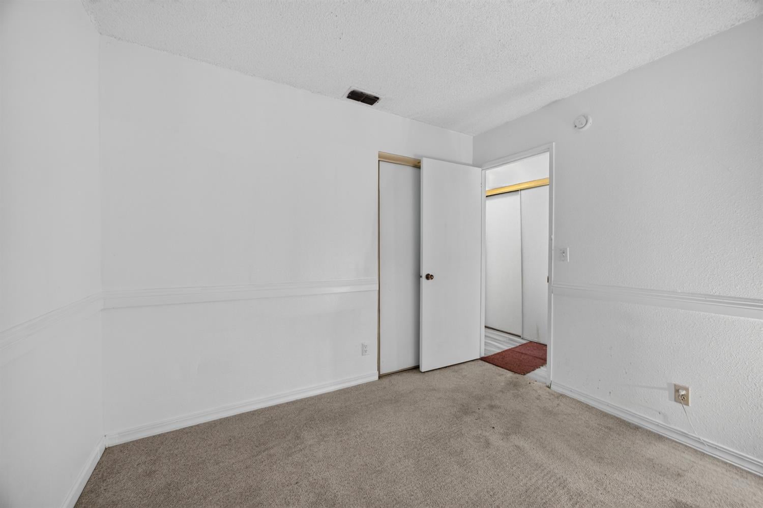 Empty bedroom with beige carpet and an open door to the closet and hallway.