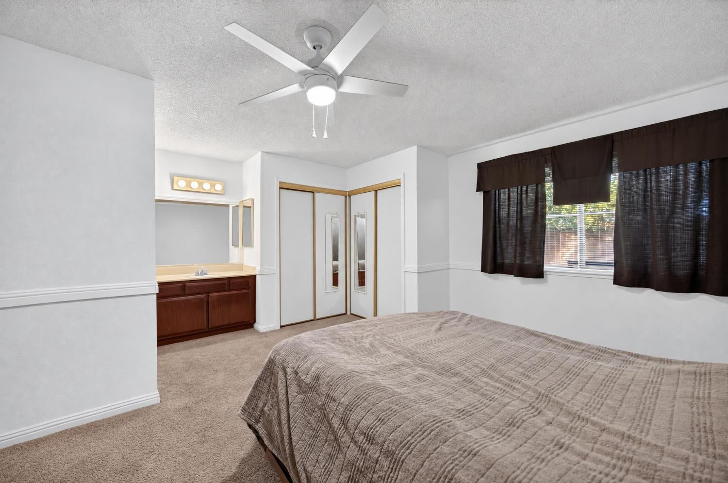 Bedroom with a large bed, ceiling fan, mirrored closet doors, and an adjoining vanity area.