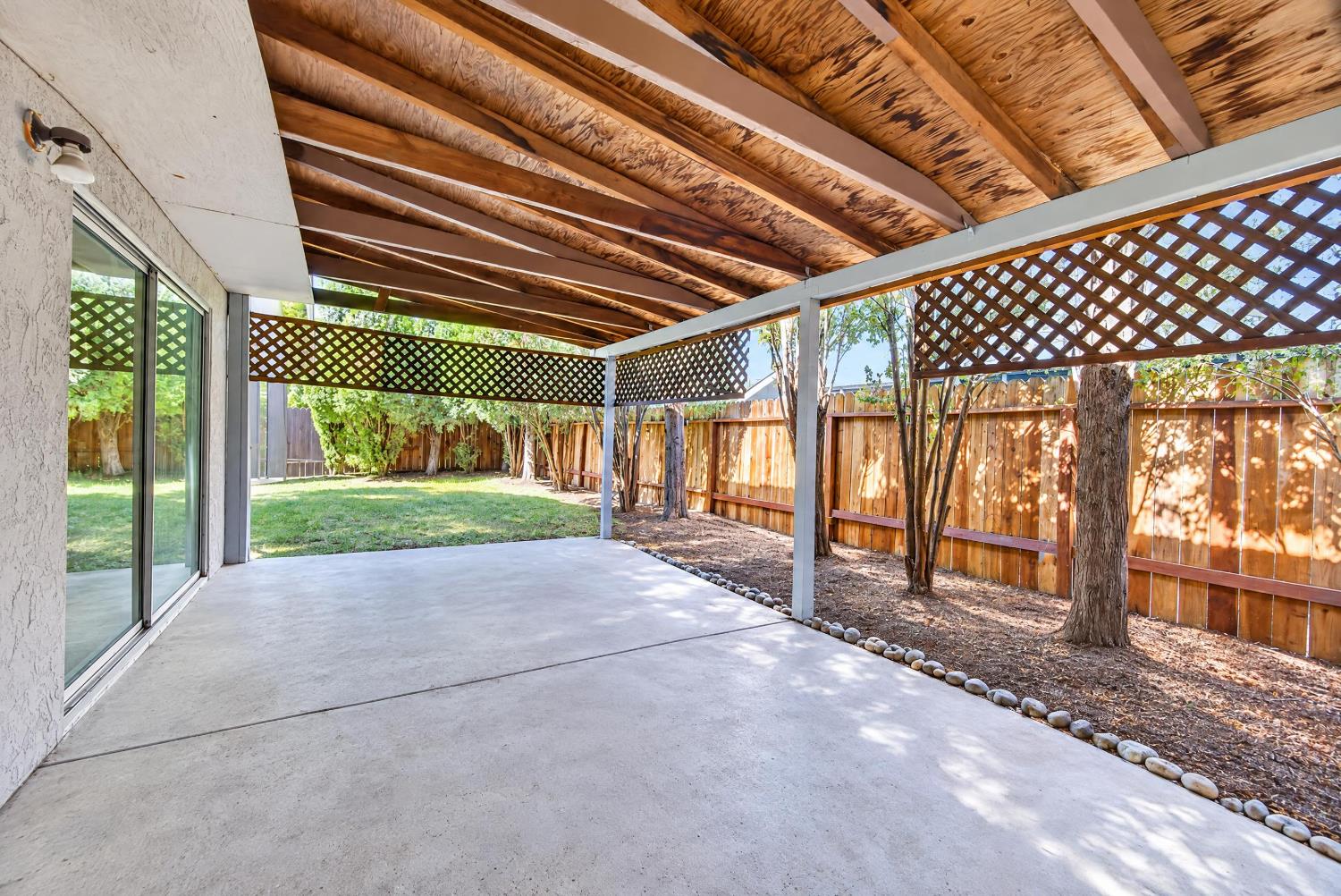 Covered concrete patio with exposed wood beams and lattice panels beside a fenced backyard.
