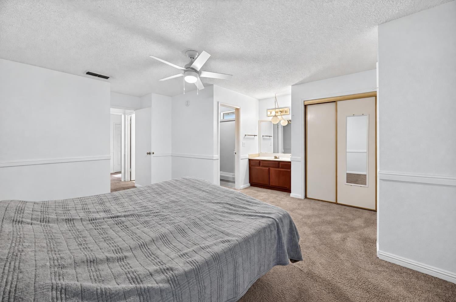 Bedroom with a gray-quilted bed, mirrored closet doors, and an adjoining bathroom vanity.
