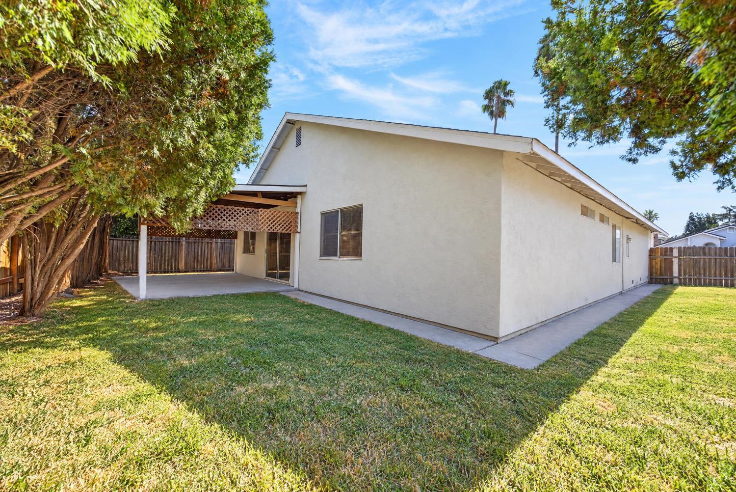 Rear of a single-story home with a covered patio and fenced, tree-shaded lawn.