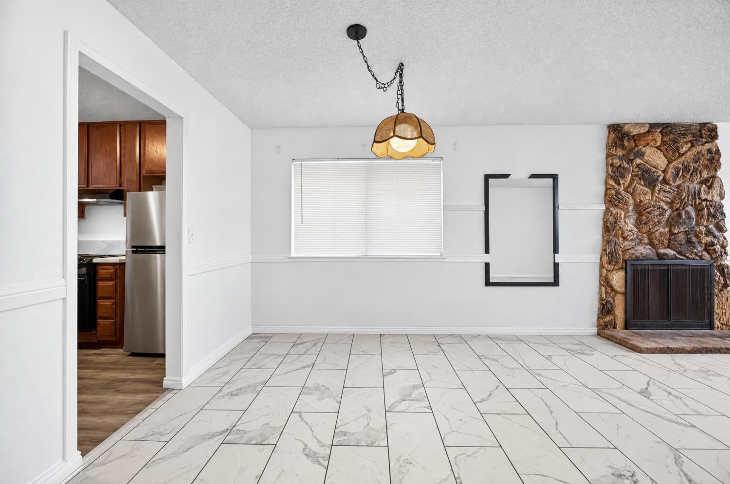 Empty dining area with marble-look tile floors, pendant light, and stone fireplace beside the kitchen.