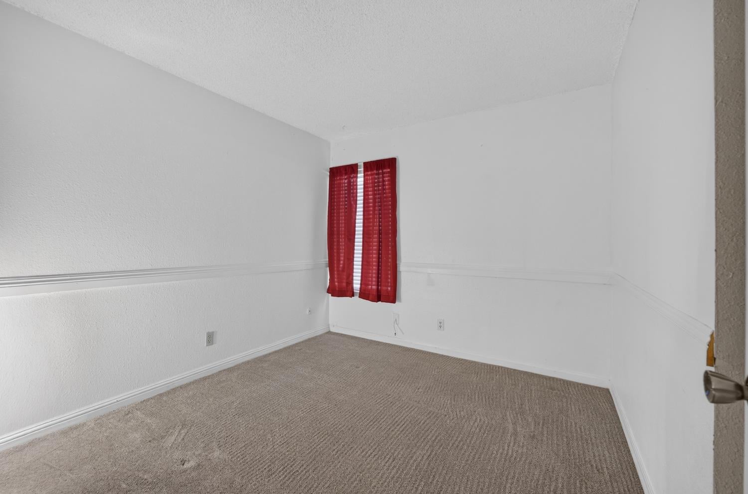 Empty bedroom with beige carpet and red curtains over a narrow window.