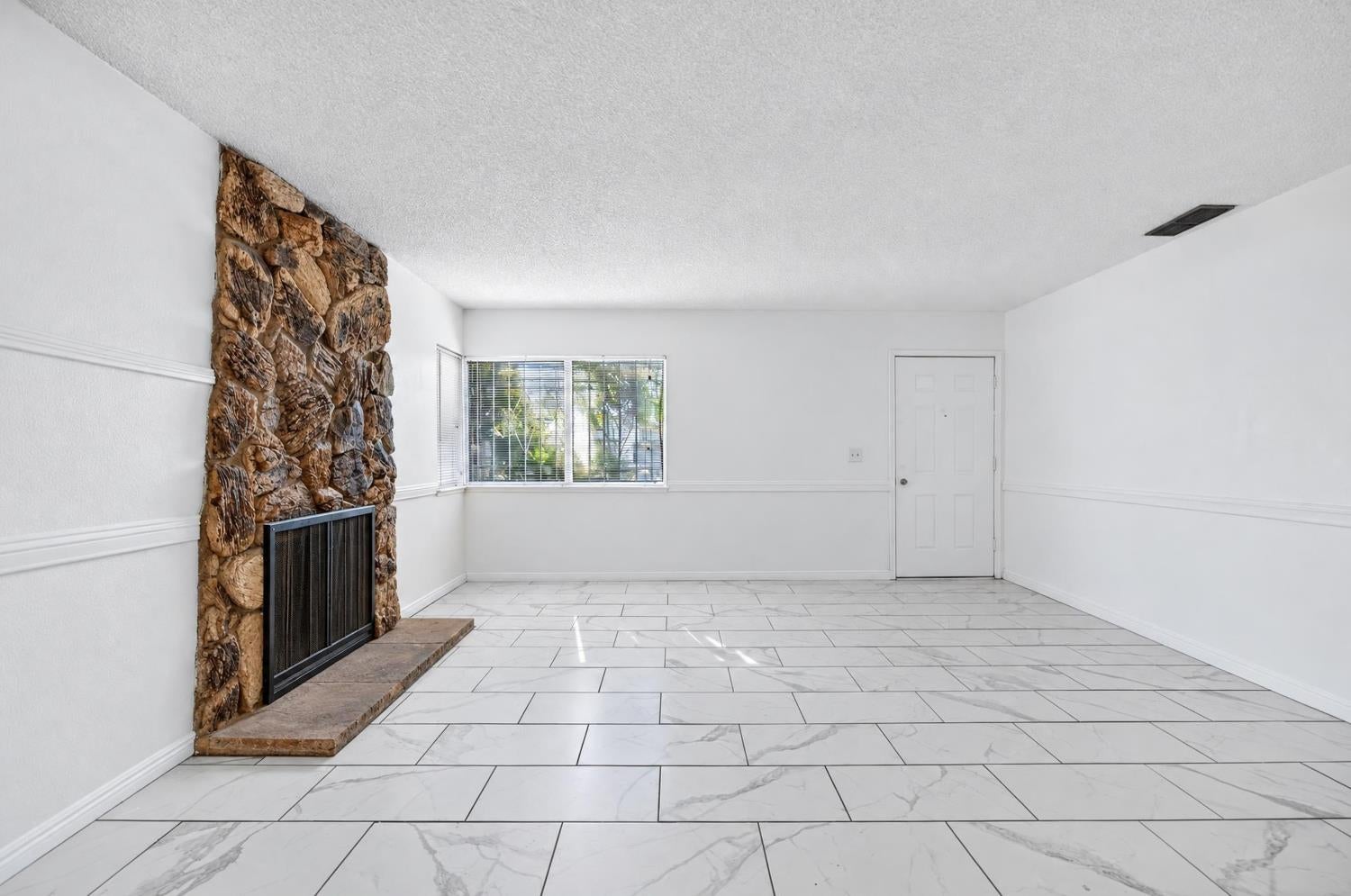 Vacant living room with a stone fireplace, large tiled floor, and wide window with blinds.
