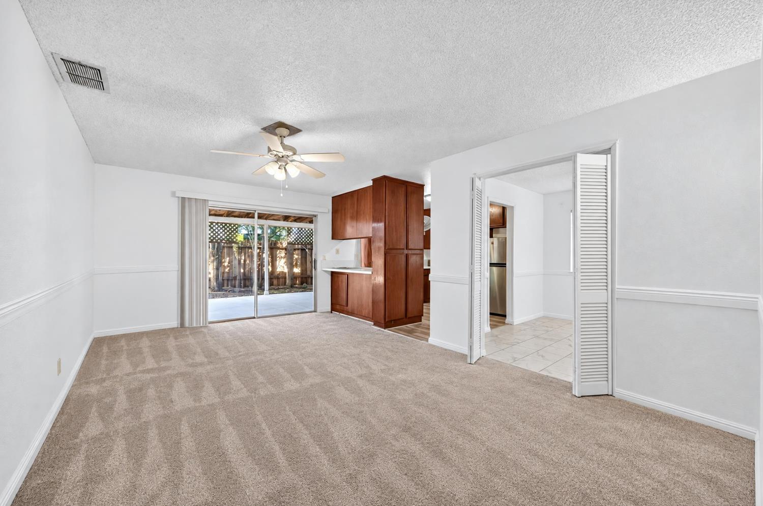 Empty carpeted living room with a ceiling fan, dark wood kitchen cabinets, and sliding patio doors.
