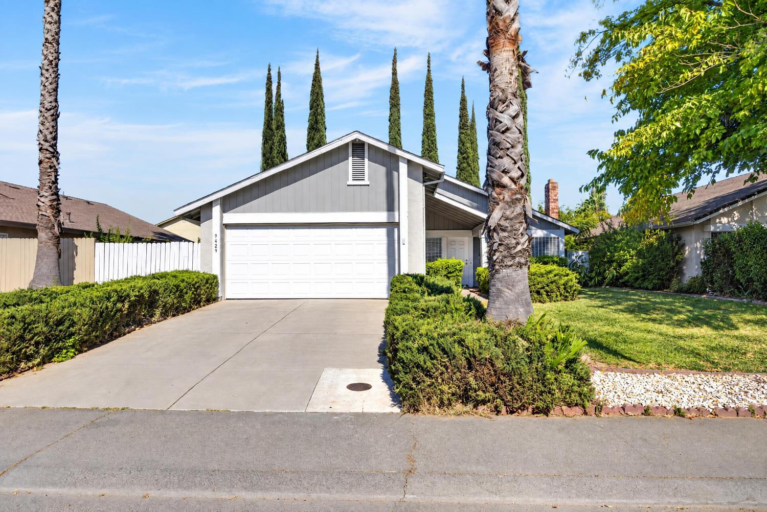 Gray single-story home with a white double garage door, broad driveway, palm trees, and trimmed landscaping.