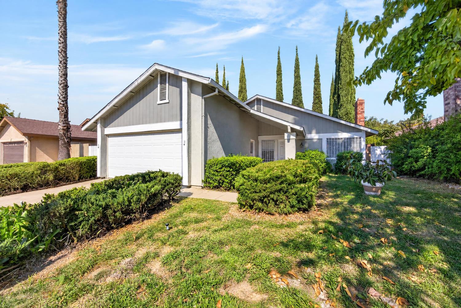 Gray-and-white single-story home with an attached garage, trimmed hedges, and tall cypress trees.