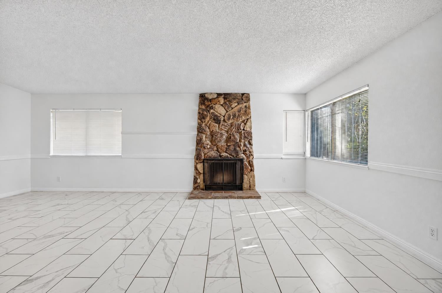 Empty living room with a rustic stone fireplace, marble-look tile floors, and wide windows.