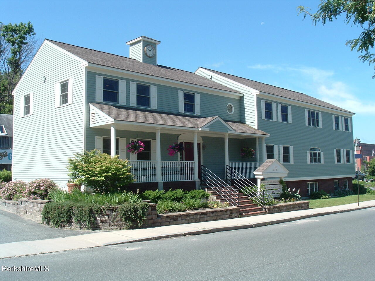 Blue-sided building with a clock tower, covered porch, flower beds, and front steps.