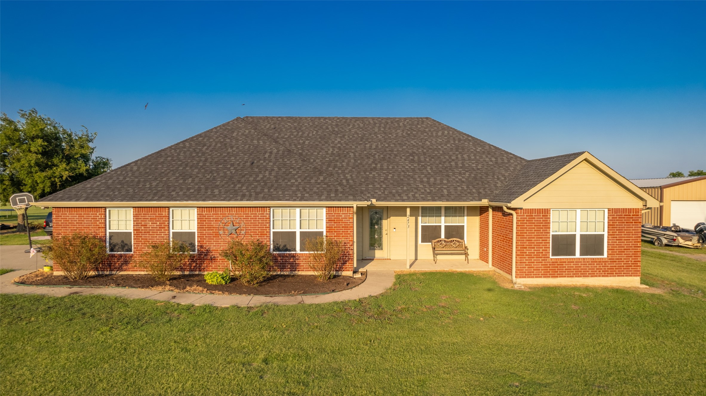 Single-story brick house with a cream-covered porch, front bench, and broad green lawn.