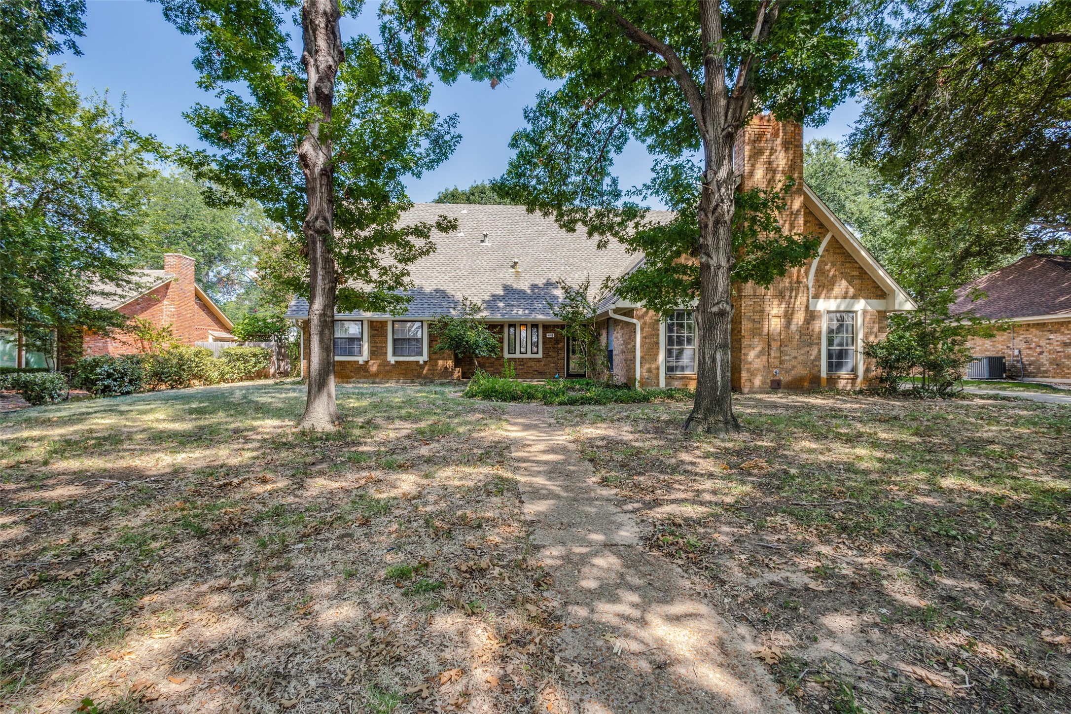 Brick house with a steep gabled roof, mature trees, and a walkway across the front lawn.