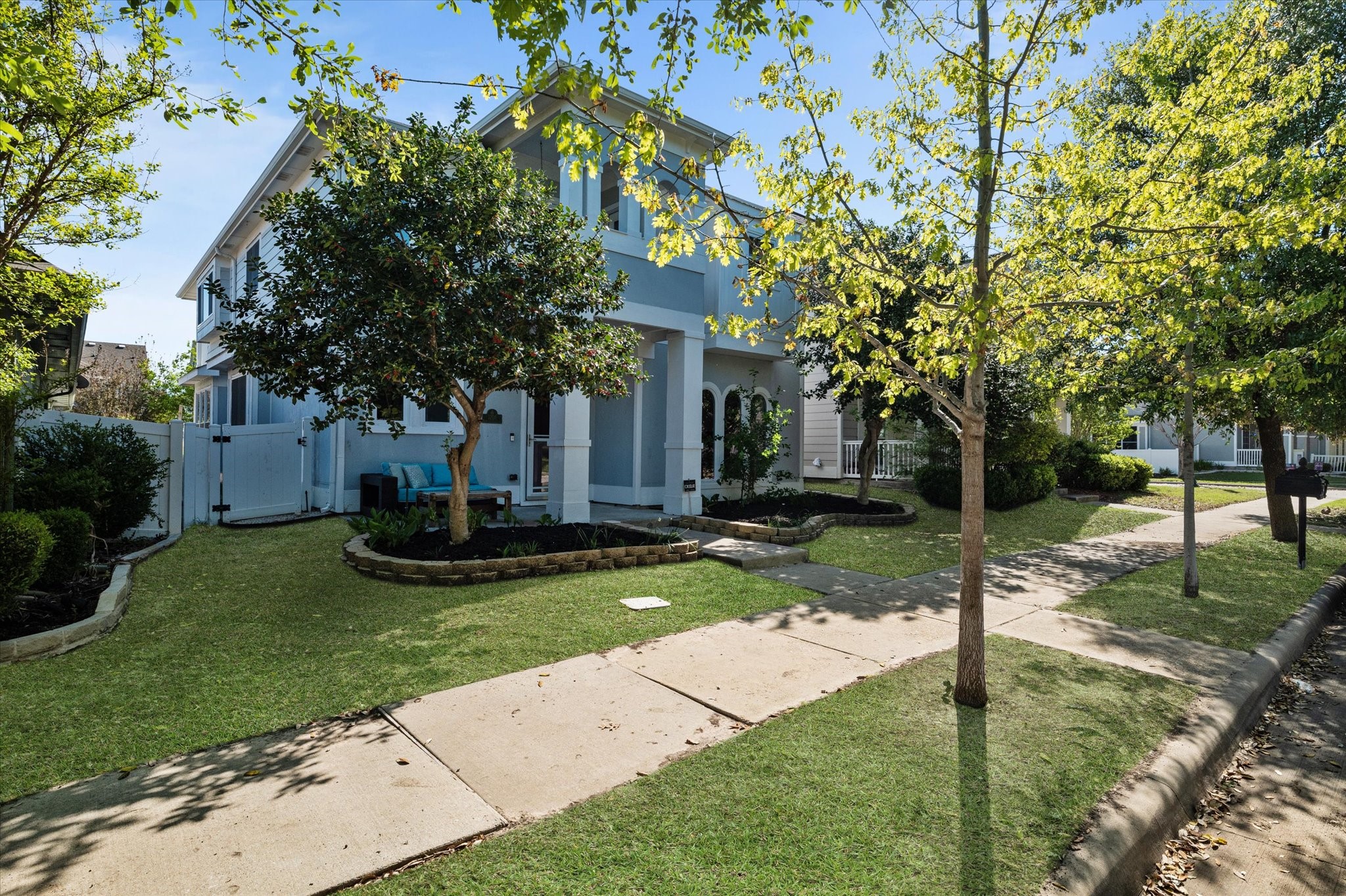 Two-story pale blue house with a covered porch, leafy trees, and a landscaped front lawn.