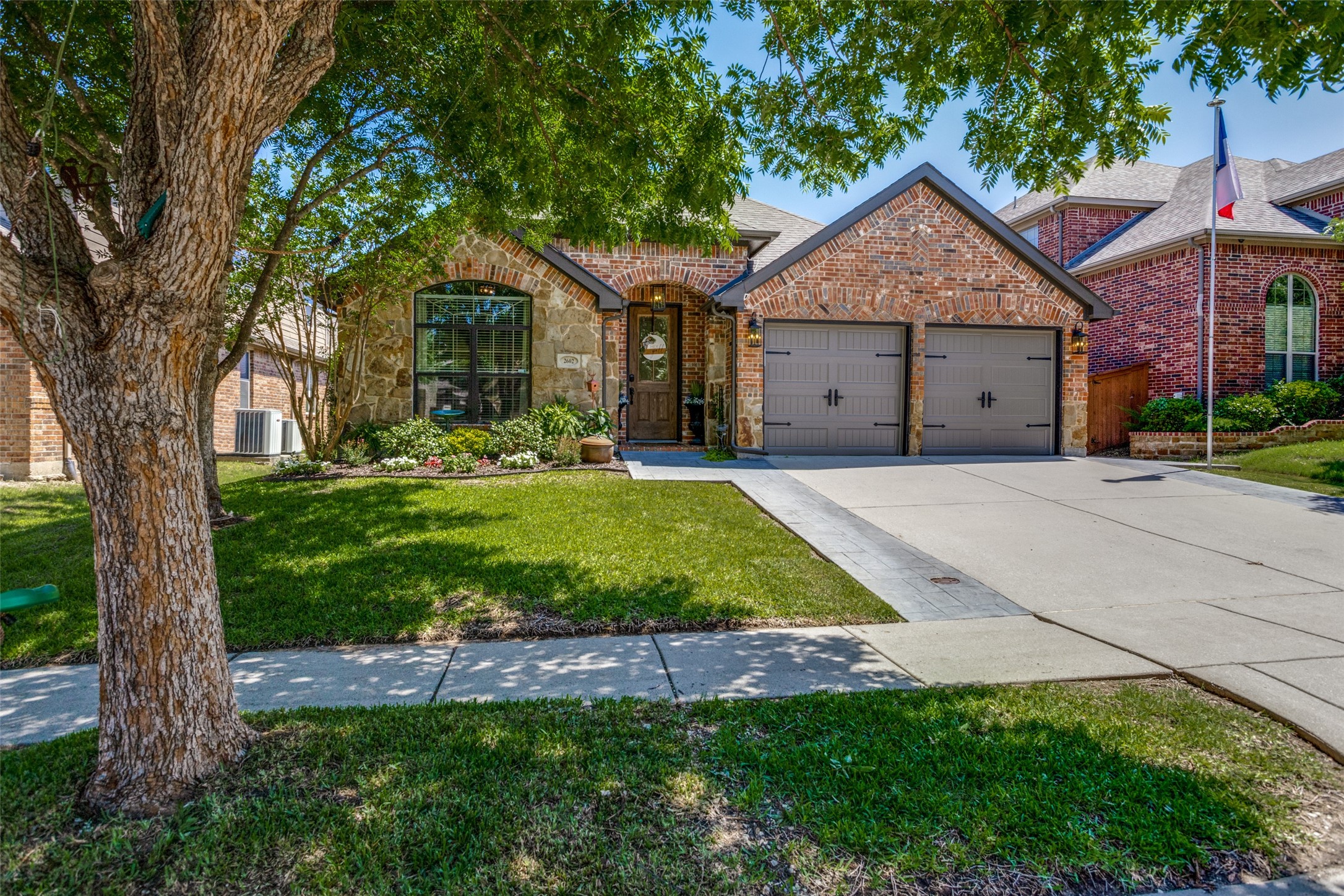 Brick-and-stone house with a double garage, shaded lawn, and mature tree in front.