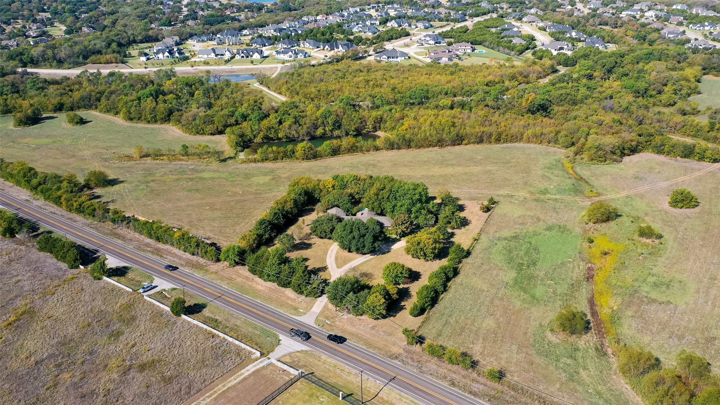 Aerial view of open acreage with a tree-shaded house, driveway, wooded creek, and road.