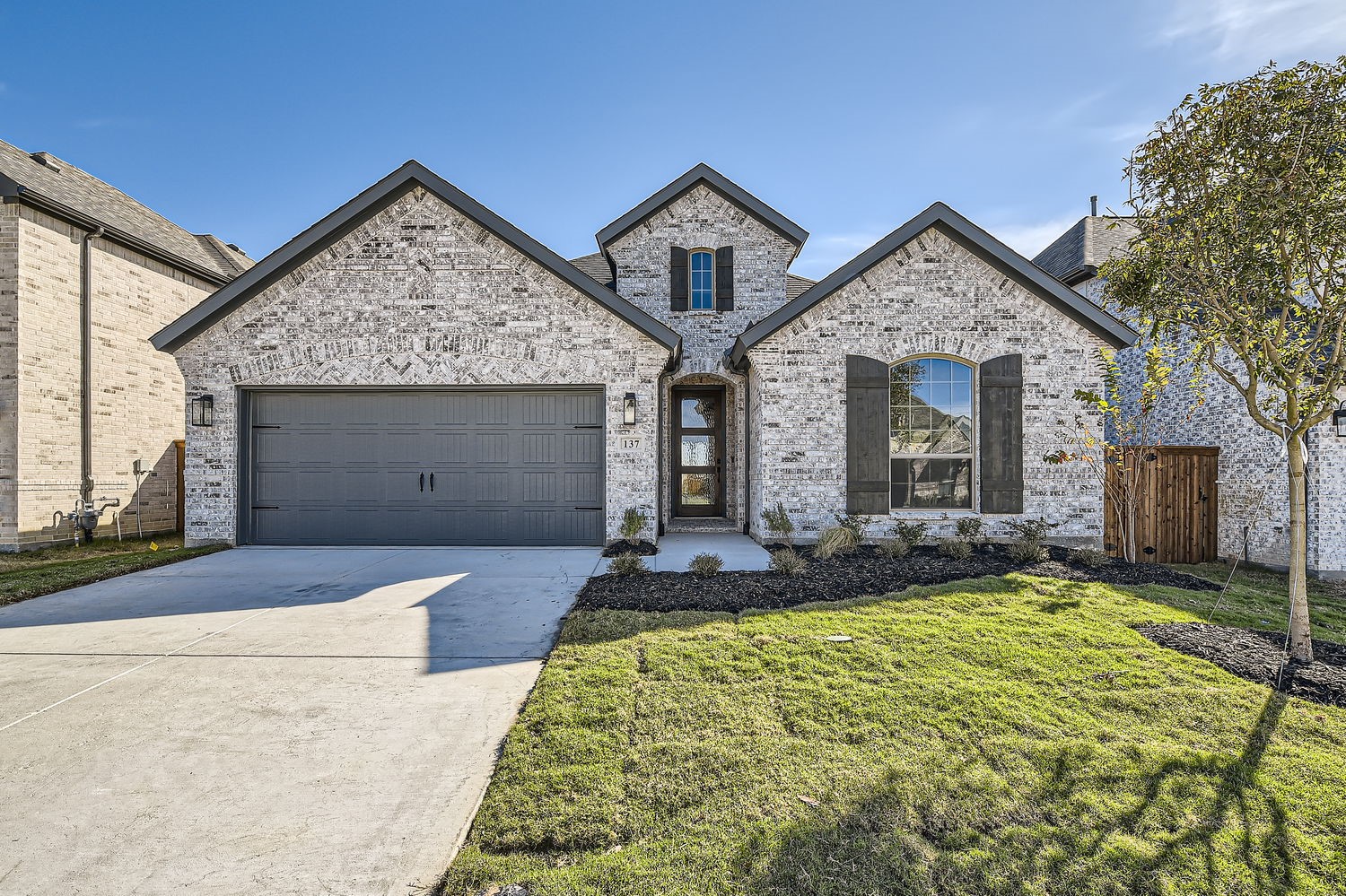 Light brick home with multiple gables, dark shutters, a two-car garage, and a landscaped front lawn.