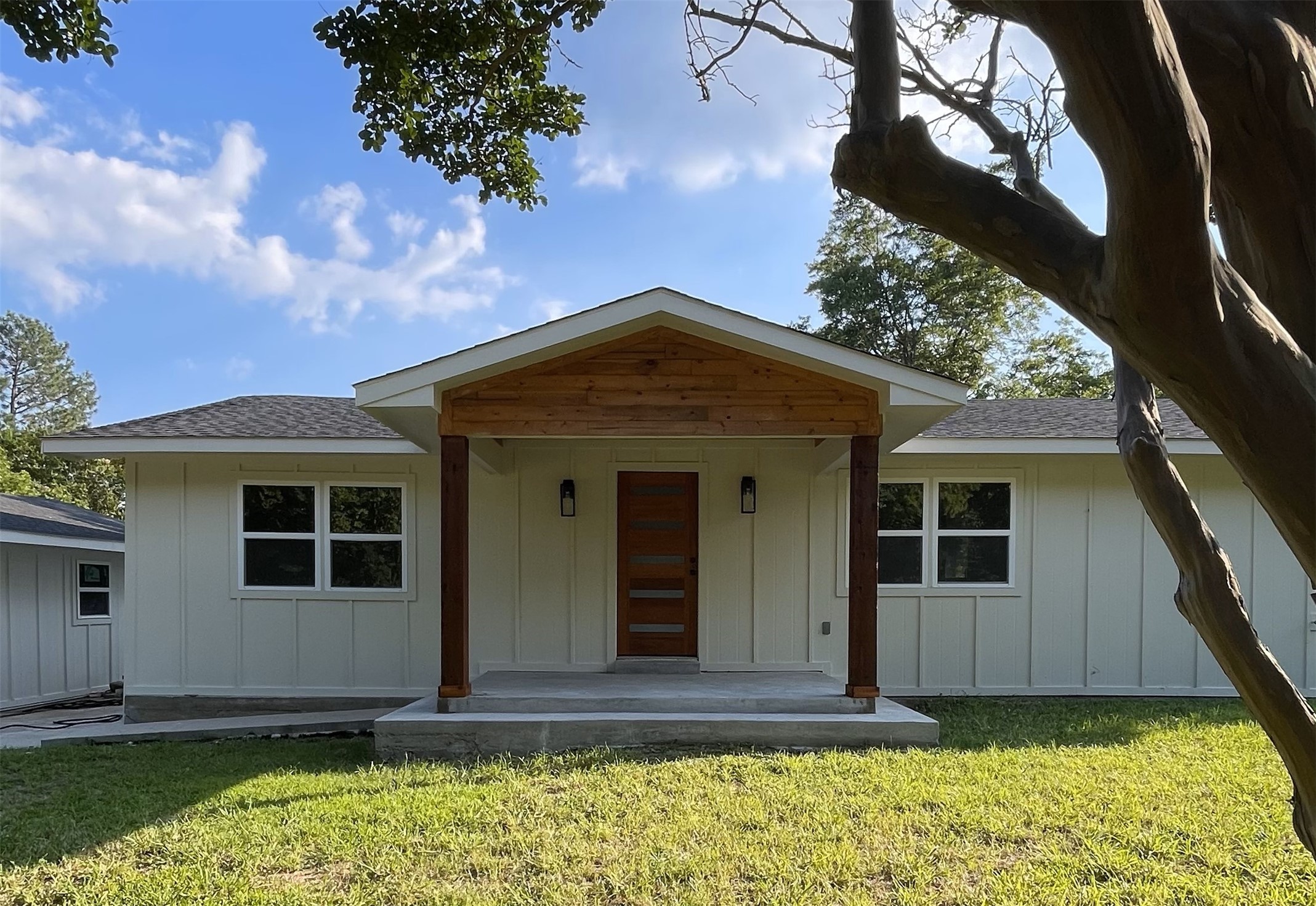 Single-story home with a wood-accented gabled porch, central wooden door, and lawn beneath mature trees.