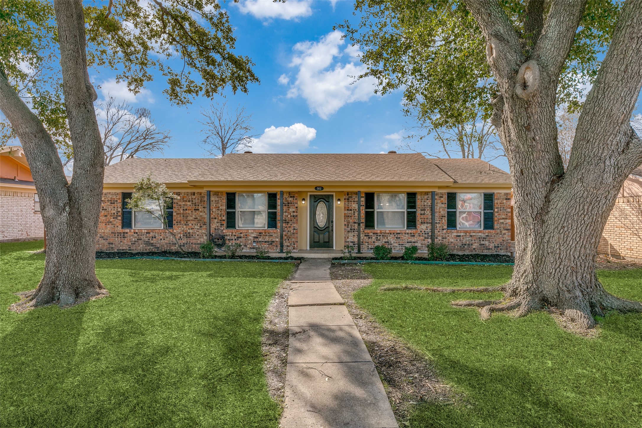 Single-story brick house framed by two large trees, with a front walkway and green lawn.