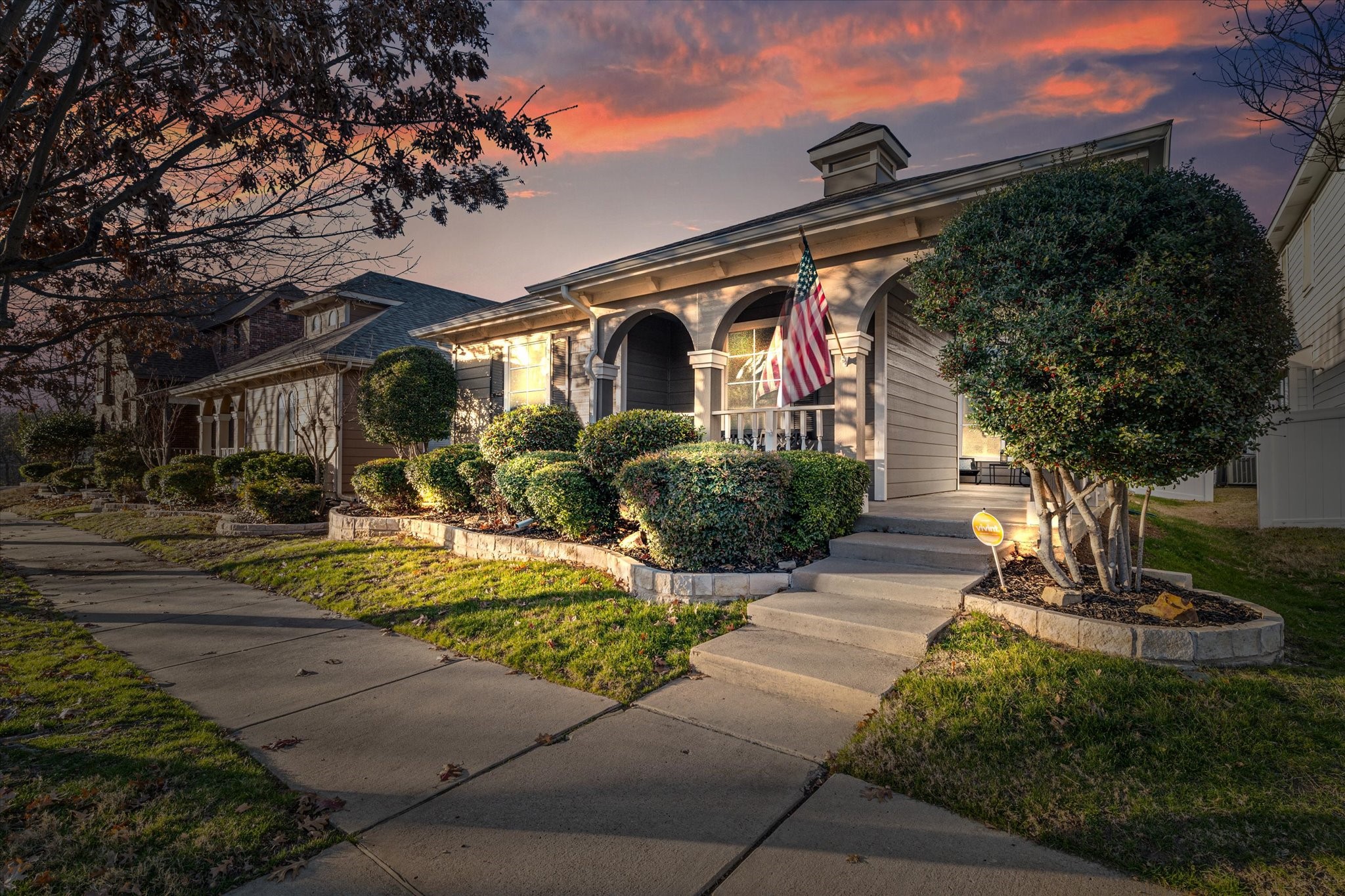 Gray house with an arched porch, American flag, and manicured shrubs at sunset.