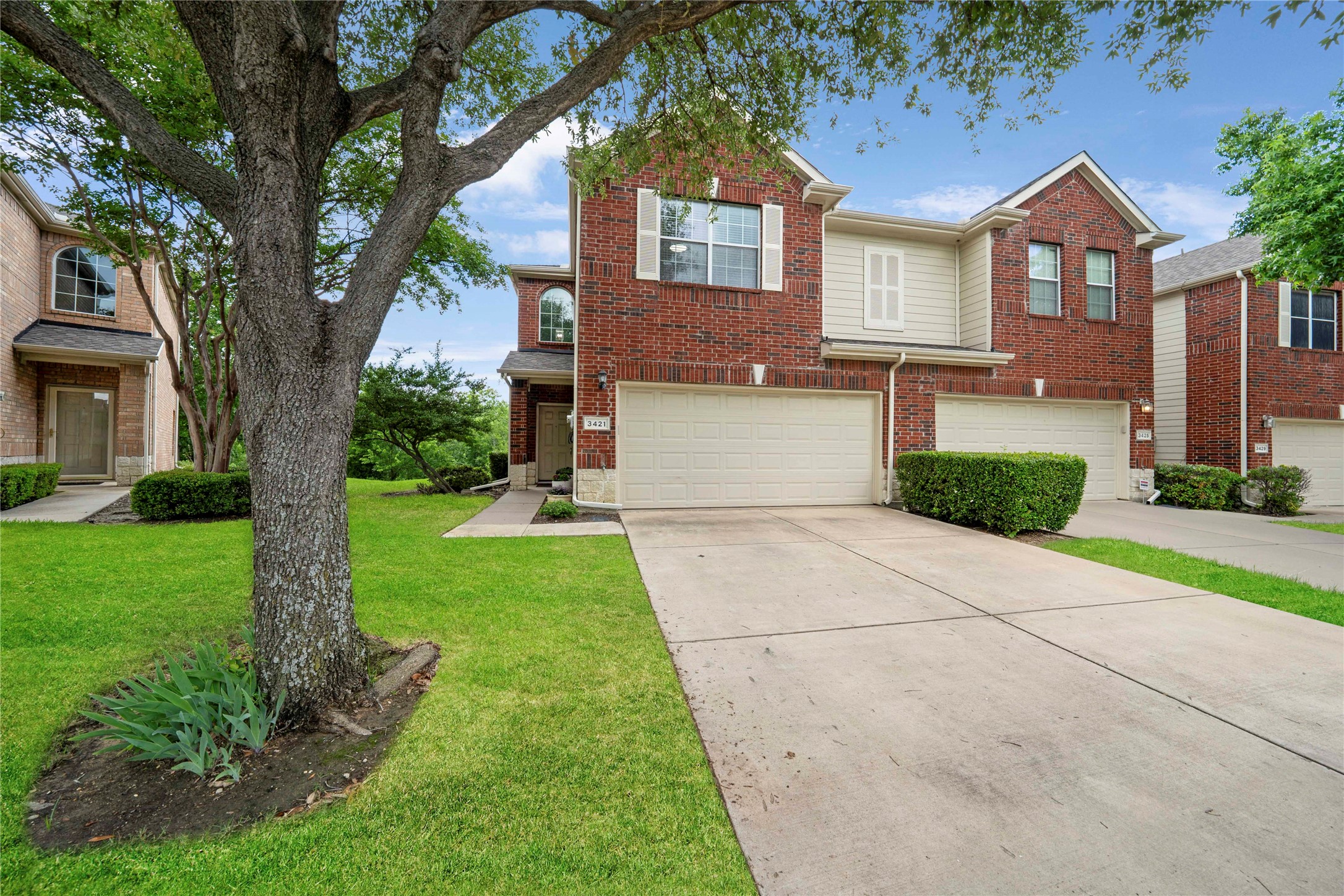 Two-story brick townhouse with a two-car garage, framed by a large shade tree and neighboring homes.